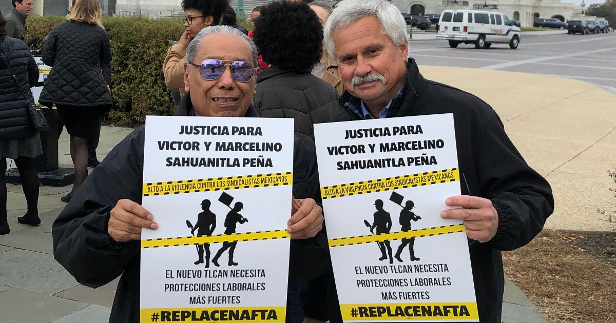 Two men holding signs that read "Justicia para Victor Y Marcelino Sahuanitla Peña. El nuevo TLCAN necesita protecciones laborales más fuertes #ReplaceNAFTA"